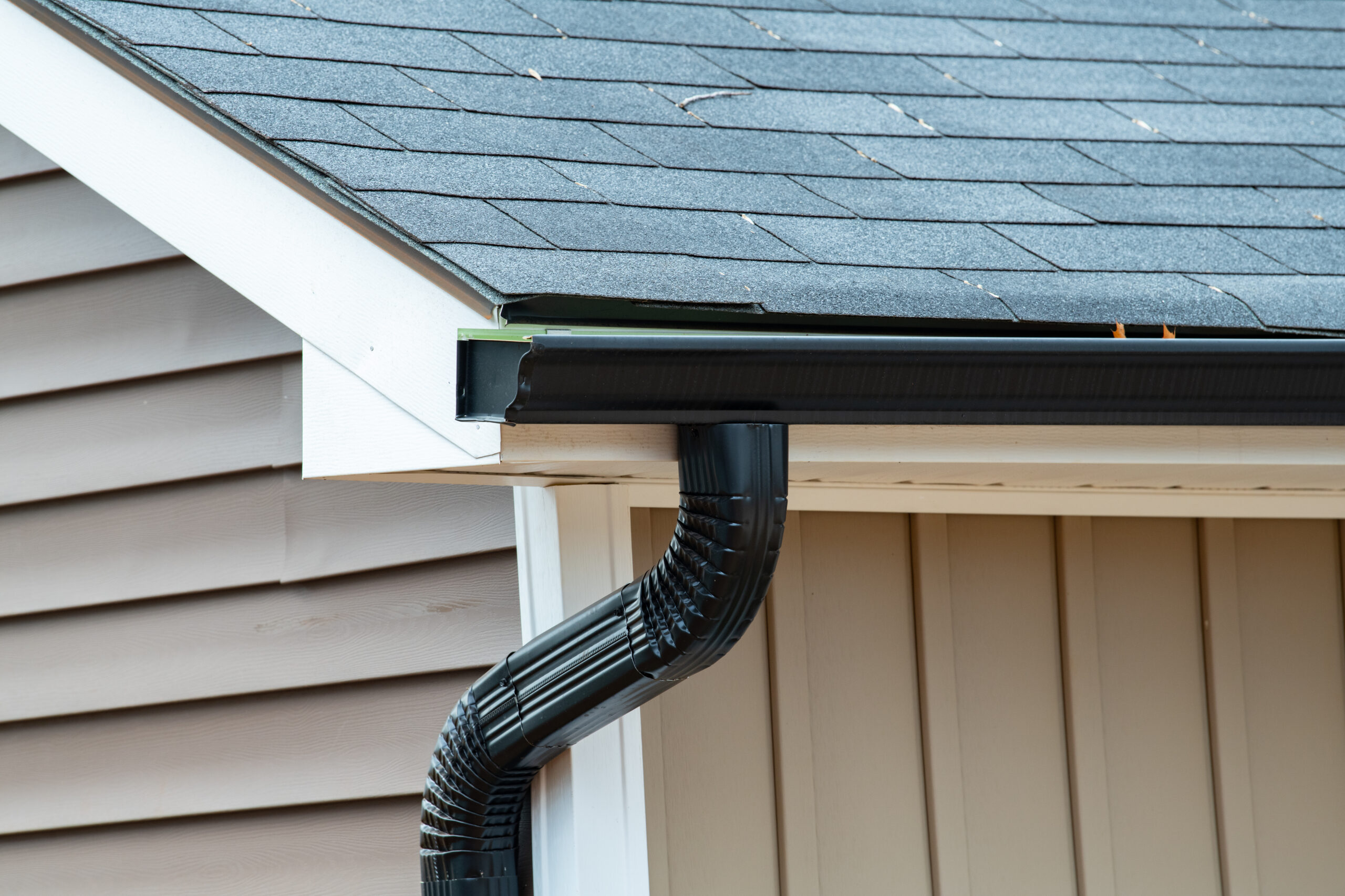 A close-up view of a gray house with black gutters, black downspout, and an asphalt shingle roof.