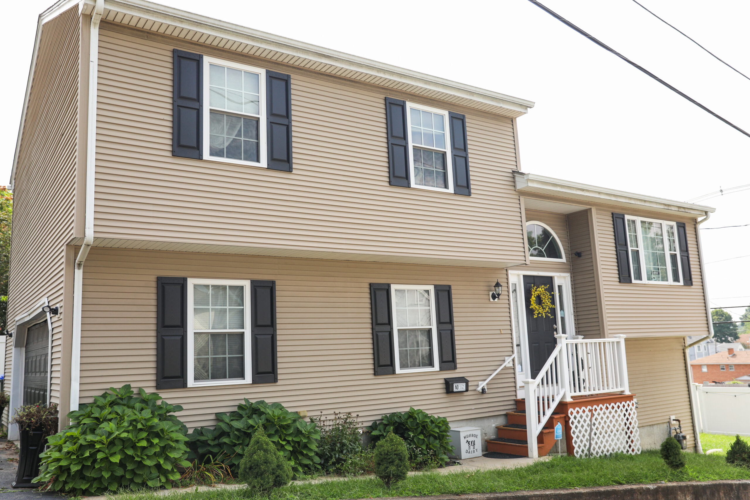 A two-story tan house with white windows, black shutters, and steps leading to the black front door with white railings.