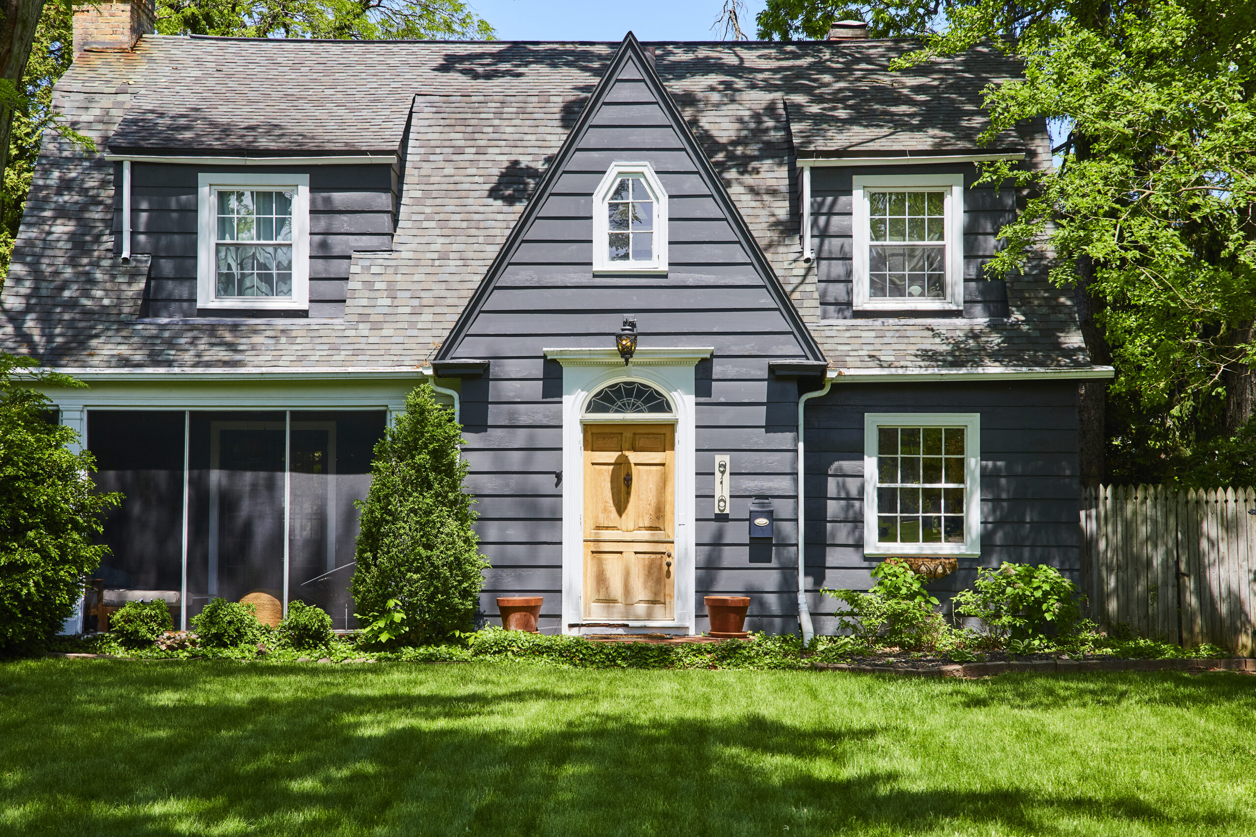 A two-story gray house with a wooden front door, white windows, and a peaked shingle roof.