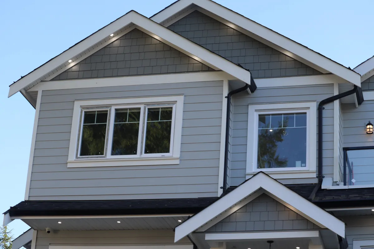 The corner of a tan house with white gutters, black windows, and an asphalt shingle roof.