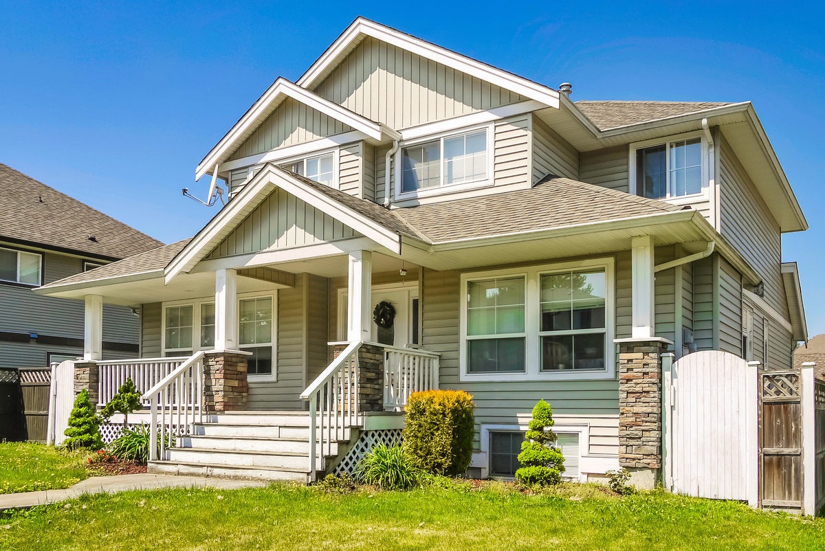A two-story tan house with a front porch, white windows, stone pillars, and an asphalt shingle roof.