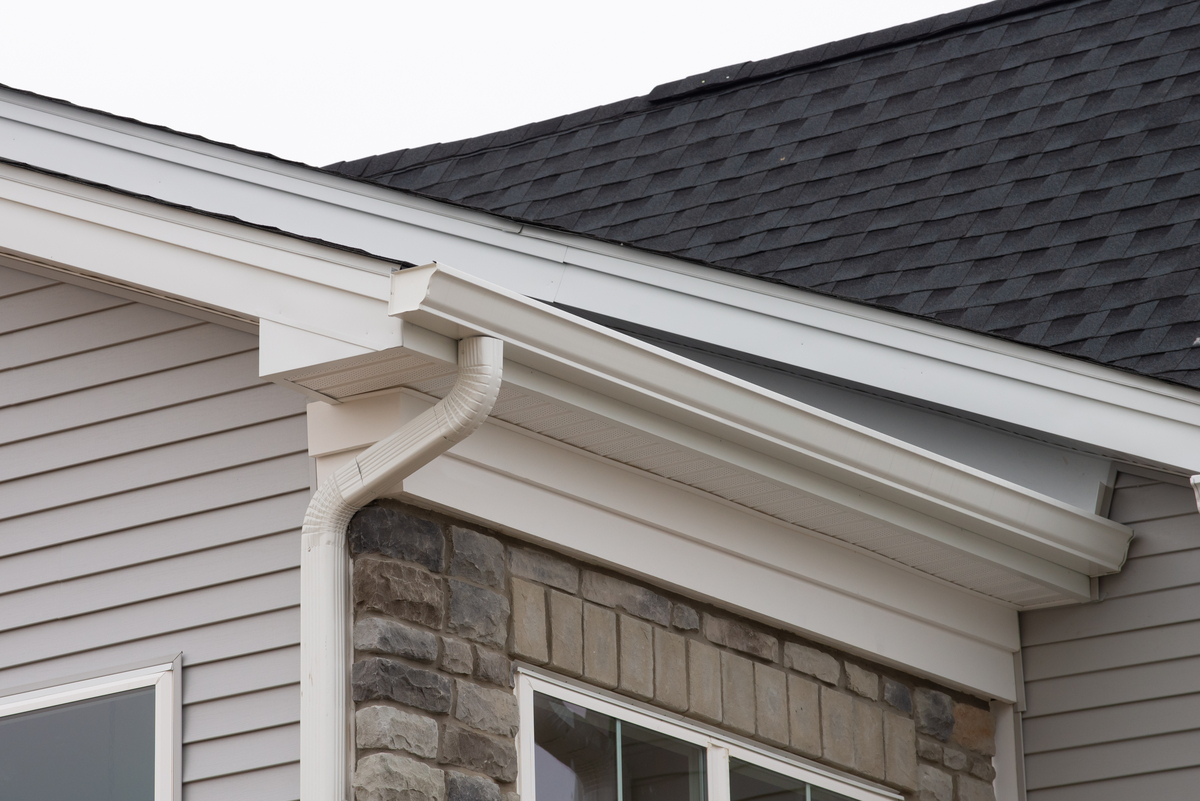 White gutters and a white downspout on a gray and stone house with an asphalt shingle roof.