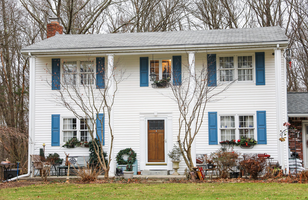A two-story white house with a wooden front door, white windows, blue shutters, and a shingle roof with a brick chimney.