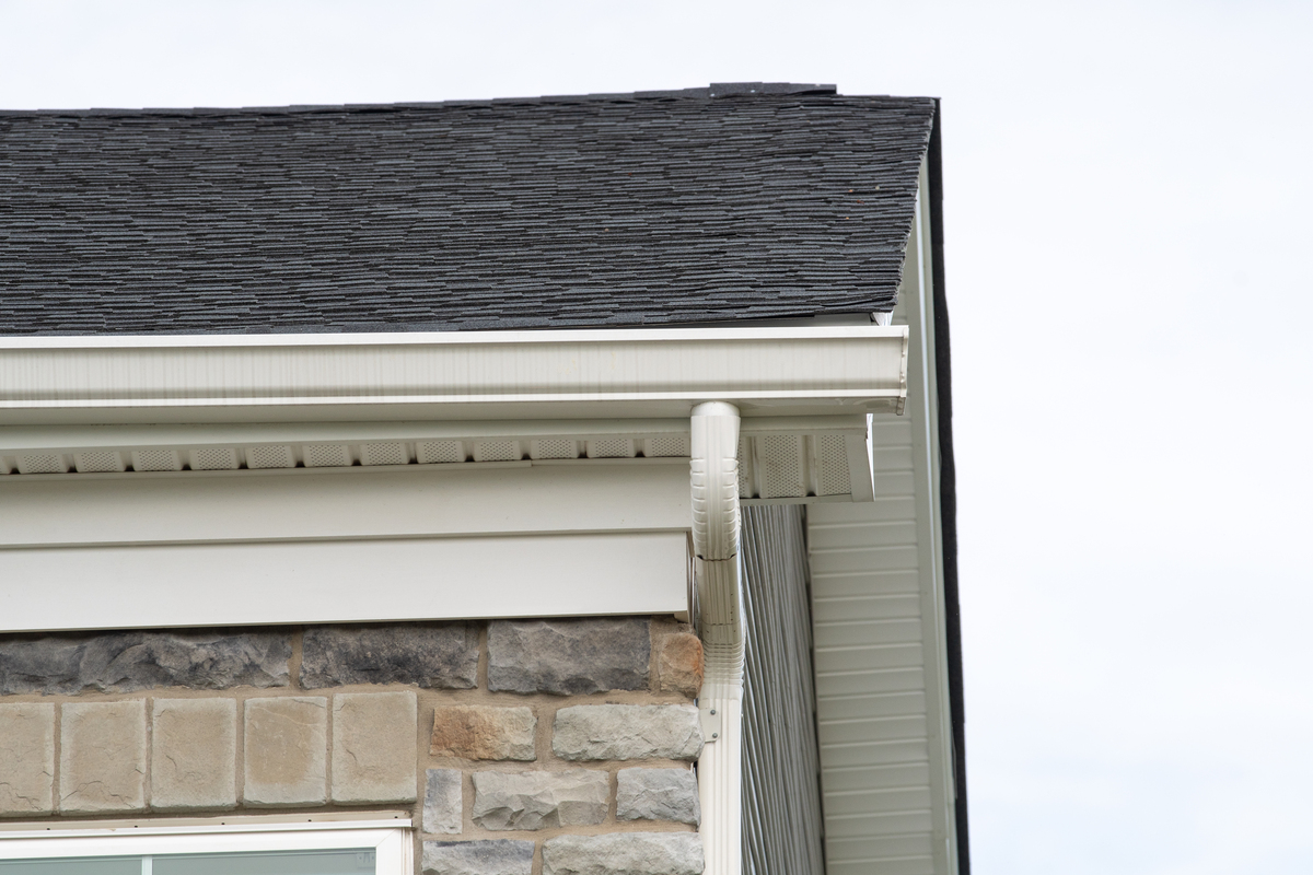 The corner of a stone house with white gutters, a white downspout, and an asphalt shingle roof.