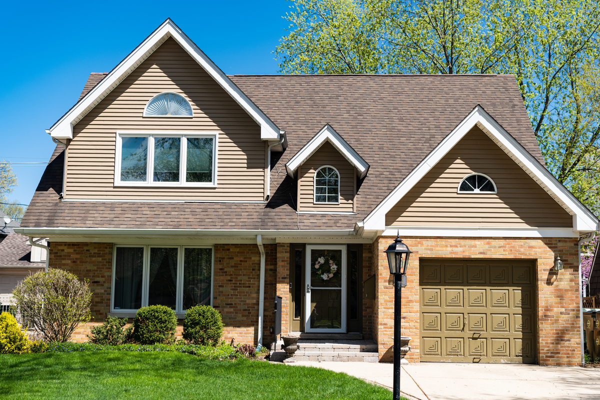 A two-story brick and tan house with a brown garage door, a wooden front door with a storm door, white windows, and an asphalt shingle roof.