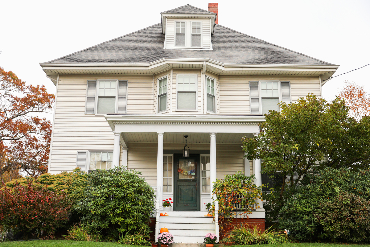 A multi-story house with a front porch, a wooden front door, white windows with gray shutters, and a shingle roof.