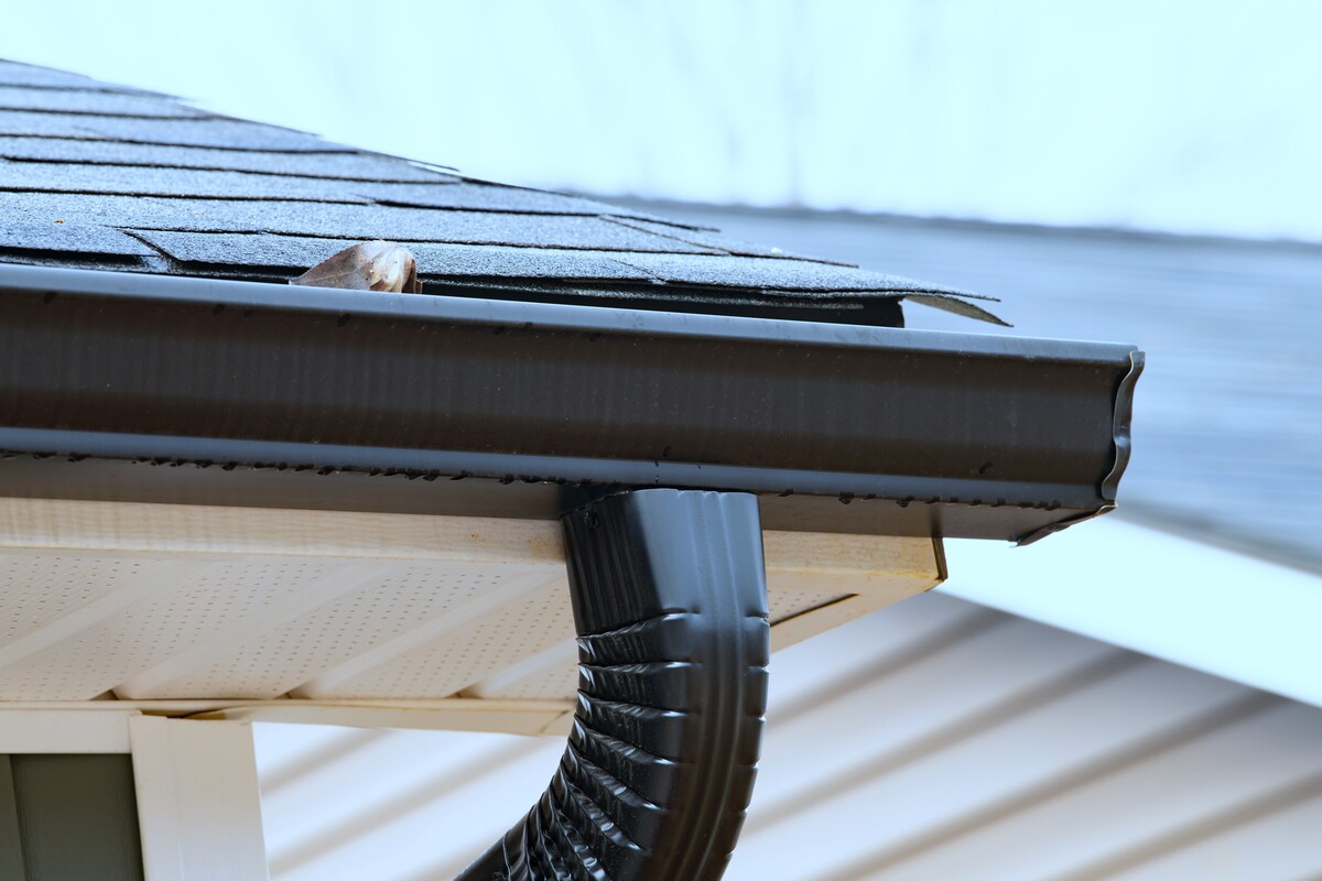 A close-up view of snow on the roof of a tan house with white windows and blue fascia.