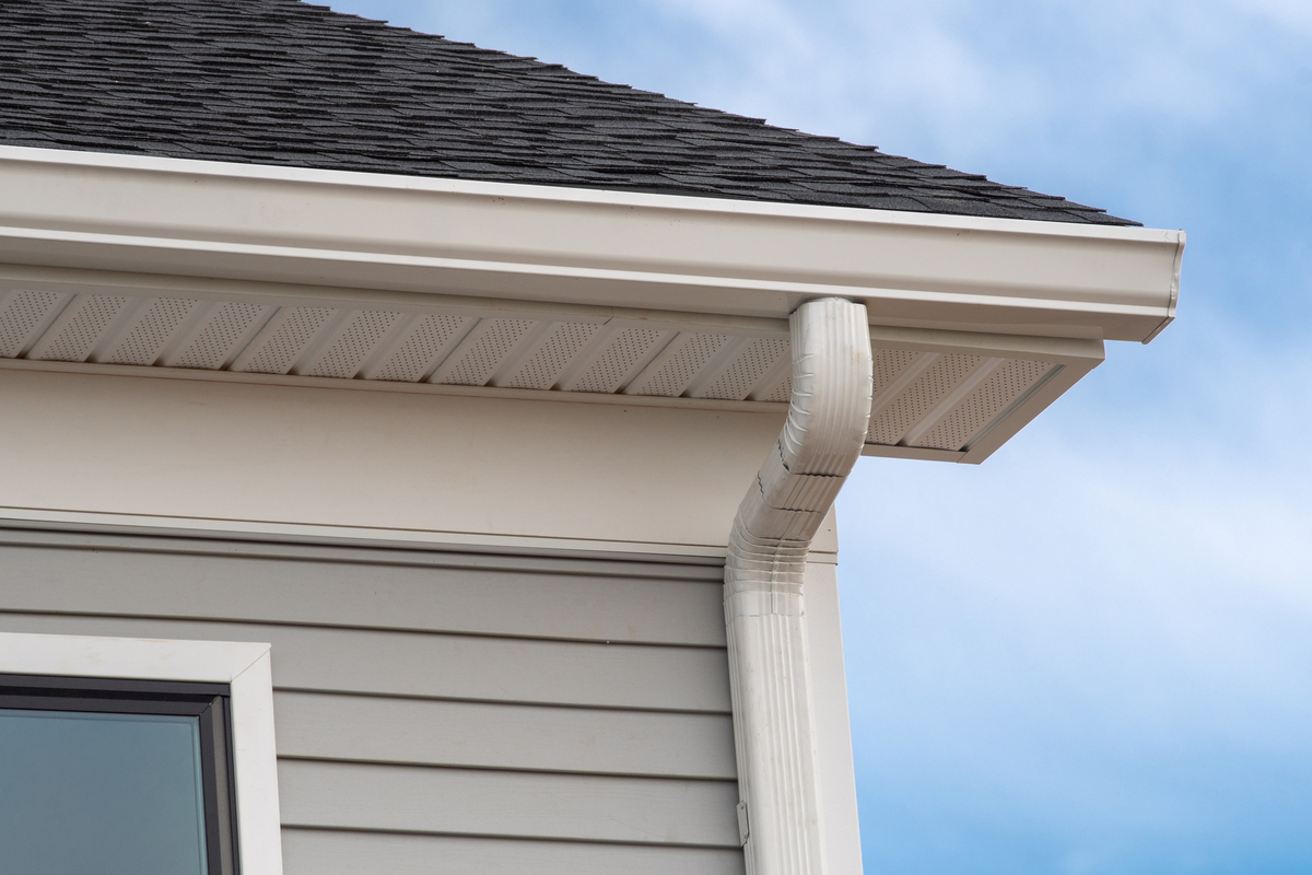 A close-up view of the corner of a gray house with white gutters, a white downspout, and an asphalt shingle roof.