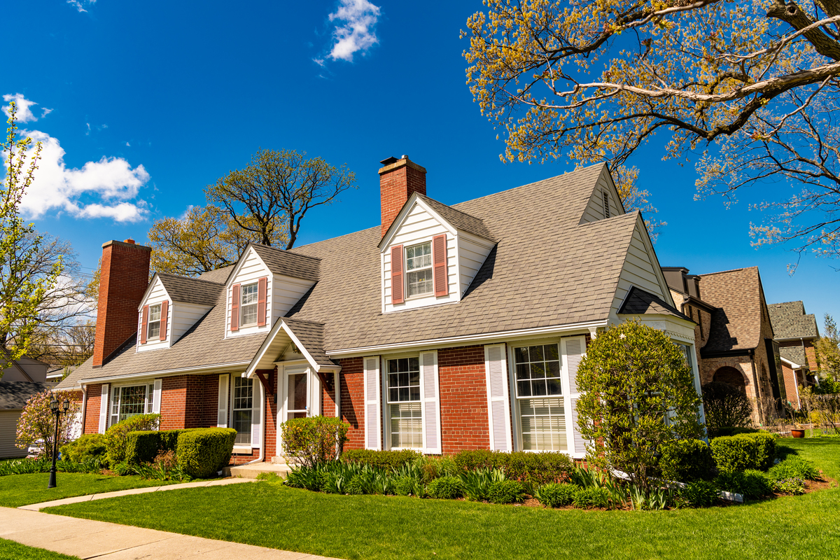 A close-up view of an asphalt shingle roof on a gray house with blue gutters and brick chimneys.