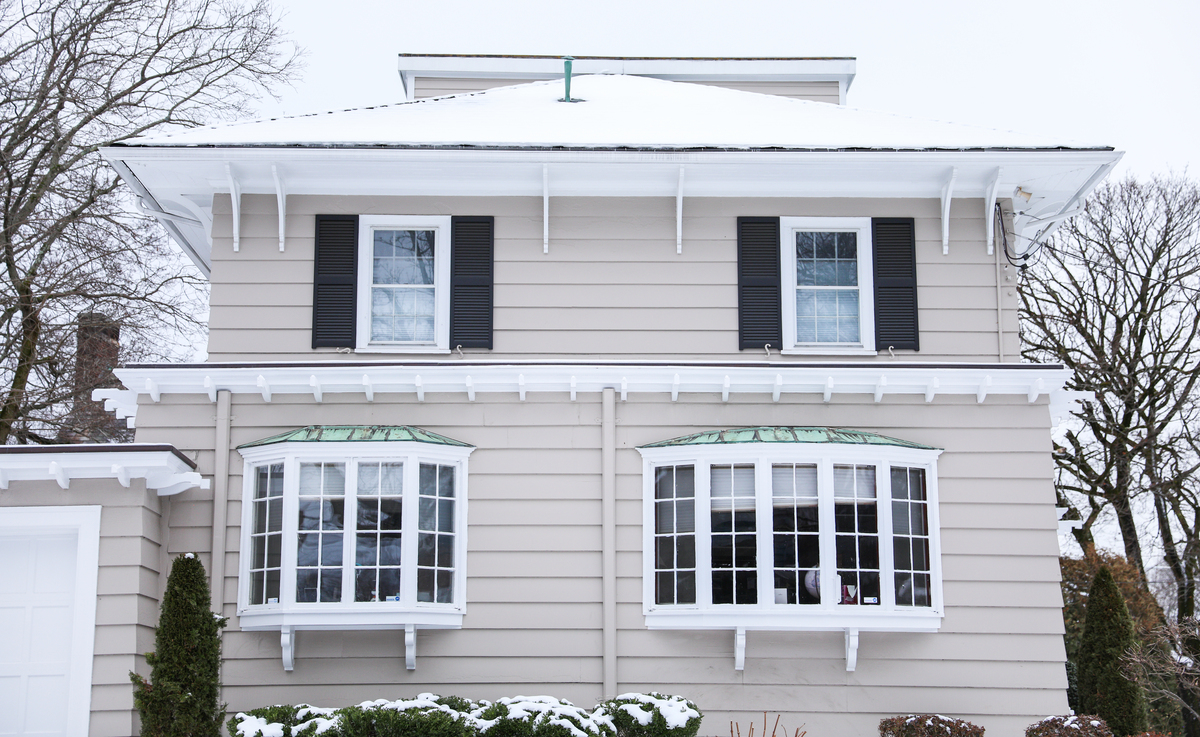 A tan house with white windows, brown shutters, and snow on the roof.